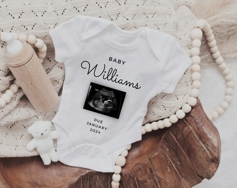 May include: White baby onesie with black text that reads "BABY Williams" and "DUE JANUARY 2024". A black and white ultrasound image is centered on the onesie.