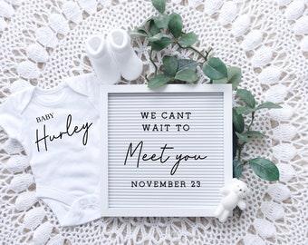 Könnte beinhalten: Ein weißer Babybody mit dem Text "Baby Hurley" in schwarzer Schrift. Ein weißes Buchstabenbrett mit dem Text "We can't wait to meet you November 23" in schwarzer Schrift. Ein weißer Teddybär und ein Paar weiße Babysocken sind ebenfalls auf dem Bild zu sehen.
