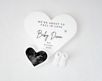 May include: White heart-shaped announcement card with the text "We're about to fall in love" and "Baby Dixon" with a small pink heart. The card also includes a due date of October 2023. A smaller heart-shaped image of an ultrasound is included, along with a pair of white baby booties.