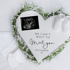 May include: White heart-shaped sign with a black and white ultrasound image, white baby socks, and a white baby hat. The sign reads "We can't wait to meet you December 23".