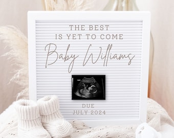 May include: White letter board with the text "The best is yet to come Baby Williams" and a black and white ultrasound image. The text "Due July 2024" is below the image. A white knitted baby bootie and a white pacifier are in the foreground.