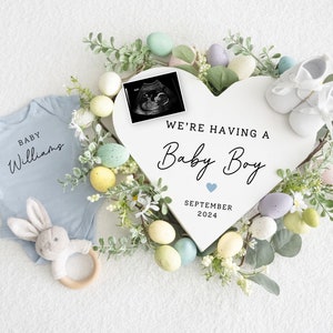 May include: A white heart-shaped sign with the text "We're having a Baby Boy" and the date "September 2024". The sign is surrounded by a wreath of green leaves and pastel-coloured eggs. A baby onesie, a pair of baby shoes, and a stuffed bunny are also included in the image.