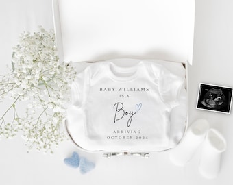 以下が含まれることがあります： 白いロンパースに「Baby Williams is a Boy」の文字と青いハートが描かれています。ロンパースは白い箱の中にあり、白い毛布、白い靴下、白黒の超音波画像が一緒に置かれています。