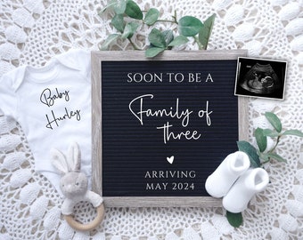 May include: A black letter board with white letters spelling "Soon to be a Family of Three" and "Arriving May 2024". A white onesie with the text "Baby Hurley" is on the left, a white bunny rattle is in the middle, and a pair of white baby socks are on the right. A black and white ultrasound image is on the letter board.