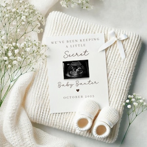 Puede incluir: Una tarjeta blanca con texto marrón que anuncia un embarazo con las palabras "We've been keeping a little secret" y una imagen de ecografía. El texto también incluye "Baby Baxter" y "October 2025". La tarjeta está sobre una manta blanca con zapatitos de bebé blancos.