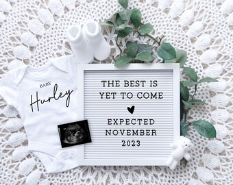 May include: White letter board with the text "The best is yet to come" and a heart symbol. Below the text is "Expected November 2023". A white onesie with the text "Baby Hurley" is in the image. A white teddy bear and a pair of white baby socks are also in the image.