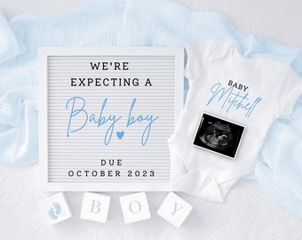 May include: A white letter board with the text "We're expecting a Baby boy" in black lettering and a blue heart. The text "Due October 2023" is also on the board. A white onesie with the text "Baby Mitchell" in blue lettering and a black and white ultrasound image is next to the letter board. Three white blocks with the letters "B", "O", and "Y" in blue lettering are below the letter board. A blue blanket is in the background.