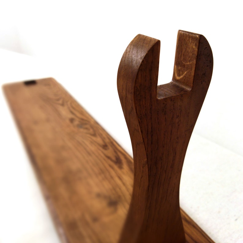 May include: A wooden sword stand with a dark brown finish. The stand features a curved support with a U-shaped top, and a long, rectangular base. The wood grain is visible, adding texture to the piece.