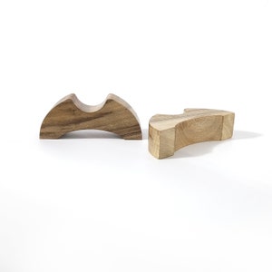 May include: Two wooden chopstick rests. One is a curved, arch-shaped design with a groove in the centre. The other is a rectangular shape with a curved edge. Both are made of light brown wood and sit on a white surface.