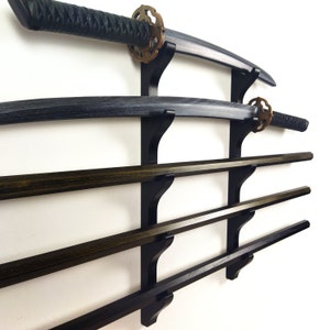Könnte beinhalten: Ein schwarzer, wandmontierter Schwerthalter mit vier Regalen. Der Halter ist aus Holz und hat eine schwarze Oberfläche. Zwei Schwerter sind auf den oberen beiden Regalen ausgestellt, und zwei Holzstöcke sind auf den unteren beiden Regalen ausgestellt.