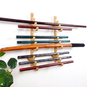 May include: A wall-mounted wooden display rack holding various wooden training weapons. The weapons are in shades of brown, red, teal, and gray. The rack is made of light-colored wood and is mounted on a white wall.