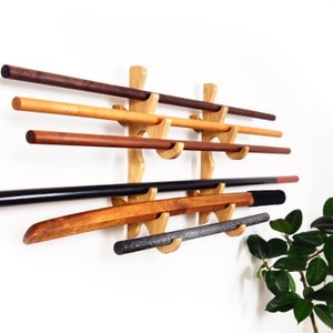 May include: A wooden wall-mounted sword rack displaying various wooden weapons. The rack is made of light-colored wood and holds several swords and staffs of different colors, including brown, black, and natural wood tones. The weapons are arranged horizontally.
