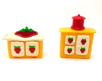 May include: Two yellow plastic toy kitchen cabinets with red strawberry details. One cabinet has a white countertop and a red sink. The other cabinet has a red lid on top.