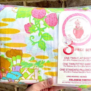 Może przedstawiać: Zestaw pościeli dla łóżka dwuosobowego Strawberry Shortcake w kolorze różowym i zielonym na białym tle. Zestaw zawiera prześcieradło płaskie, prześcieradło z gumką i poszewkę na poduszkę. Opakowanie zawiera tekst "Witamy w świecie Strawberry Shortcake" i "Zestaw 5-częściowy".