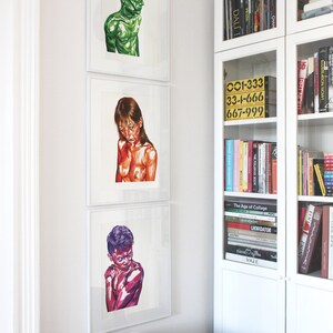 May include: Three framed illustrations of people, each with a different color scheme. The first illustration is of a person with blue hair and a green body. The second illustration is of a person with brown hair and a red body. The third illustration is of a person with purple hair and a pink body. The illustrations are hanging on a white wall next to a white bookshelf filled with books.