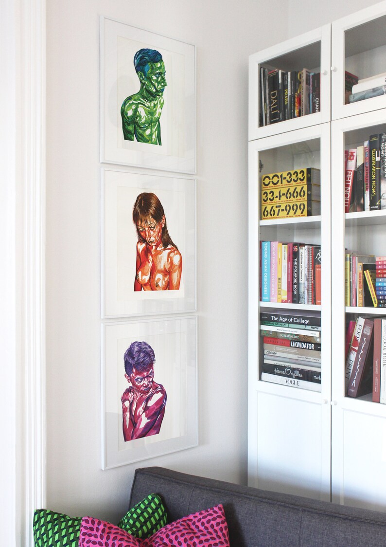 May include: Three framed illustrations of people, each with a different color scheme. The first illustration is of a person with blue hair and a green body. The second illustration is of a person with brown hair and a red body. The third illustration is of a person with purple hair and a pink body. The illustrations are hanging on a white wall next to a white bookshelf filled with books.