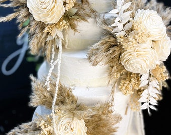 May include: A tiered white wedding cake decorated with cream-colored roses, dried pampas grass, and delicate white foliage. The cake has a textured finish and is adorned with floral arrangements. The overall design is elegant and rustic.
