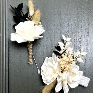 May include: Two white sola flower boutonnieres with dried grasses and greenery. One boutonniere has a black pin and the other has a white elastic band.