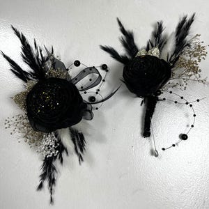 May include: Two black floral boutonnieres with feathers, glitter, and decorative accents. One boutonniere has a stem, while the other has a decorative ribbon and bead design. The boutonnieres are likely for a wedding or formal event.