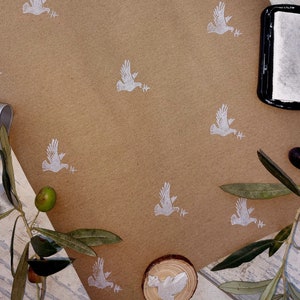 May include: Brown kraft paper featuring a repeating pattern of white doves holding olive branches. The scene includes olive branches, olives, gold scissors, a silver ribbon, and a black ink pad, ideal for crafts or stationery.