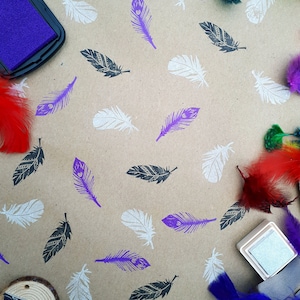 May include: A brown paper sheet with various feather stamps in black, white, and purple. There are also real feathers in red, blue, and purple. The image includes two ink pads, one purple and one silver.