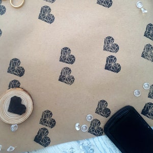 May include: Brown craft paper with black geometric heart stamps. The paper is surrounded by scissors, black ribbon, and clear plastic gems.