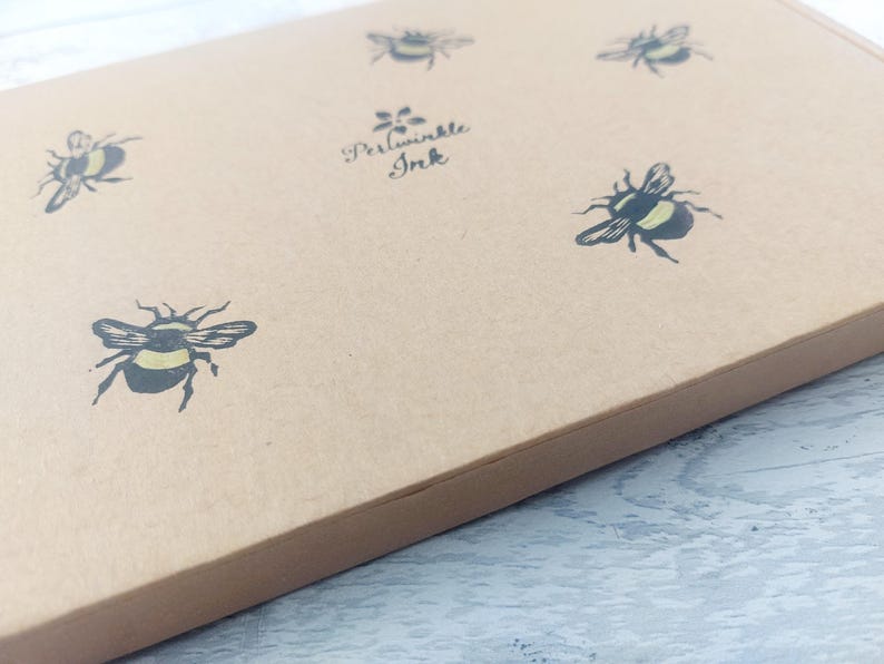 May include: A light brown box with a repeating pattern of black and yellow bumblebees. The text "Periwinkle Ink" is printed in black script. The box is angled, showing the top and side.