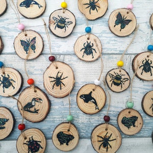 May include: A collection of wooden ornaments featuring a variety of nature-themed designs, including butterflies, bees, spiders, beetles, turtles, and a toucan. The ornaments are painted in black and white and have a rustic, natural look.