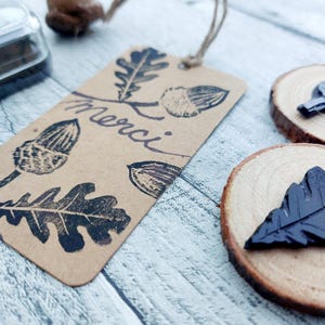 May include: A brown paper tag with black oak leaf and acorn stamps and the word "merci" in cursive. Two wooden slices with black leaf stamps are also visible. The tag is attached with a natural twine string.