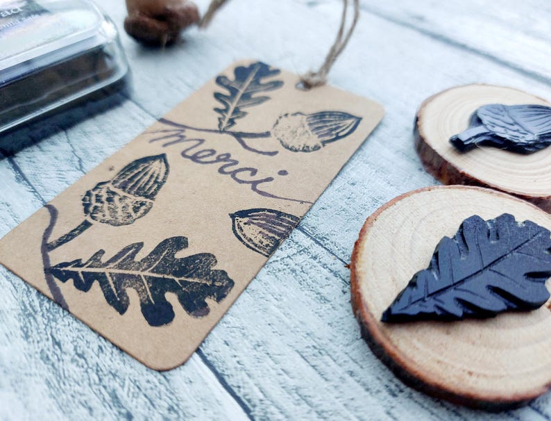 May include: A brown paper tag with black oak leaf and acorn stamps and the word "merci" in cursive. Two wooden slices with black leaf stamps are also visible. The tag is attached with a natural twine string.