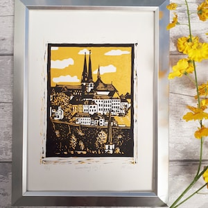 May include: A framed black and white linocut print of a cityscape with two tall church spires. The print is on a yellow background with white clouds. The title of the print is "Leerlingen City" and the date is "18.2021".