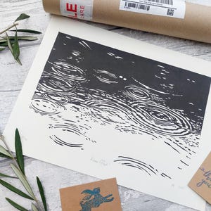 May include: Black and white art print depicting a rain scene with concentric circles. The print is on white paper and is accompanied by a brown cardboard tube, a small card with a feather design, and a small card with a rabbit design.