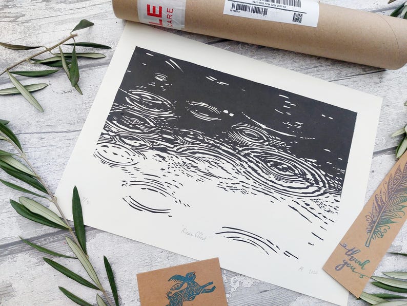 May include: Black and white art print depicting a rain scene with concentric circles. The print is on white paper and is accompanied by a brown cardboard tube, a small card with a feather design, and a small card with a rabbit design.