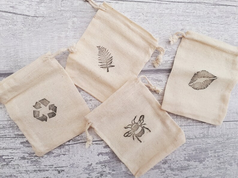 May include: Four small, white cotton drawstring bags with different designs printed on them. The designs include a fern leaf, a bee, a recycle symbol, and a pair of lips.