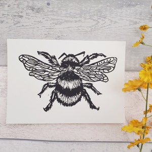 Puede incluir: Una tarjeta rectangular blanca que presenta una impresi&oacute;n en bloque de tinta negra de un abejorro. La abeja est&aacute; representada con alas detalladas y estampadas, y un cuerpo difuso y texturizado. La ilustraci&oacute;n muestra l&iacute;neas negras de alto contraste.