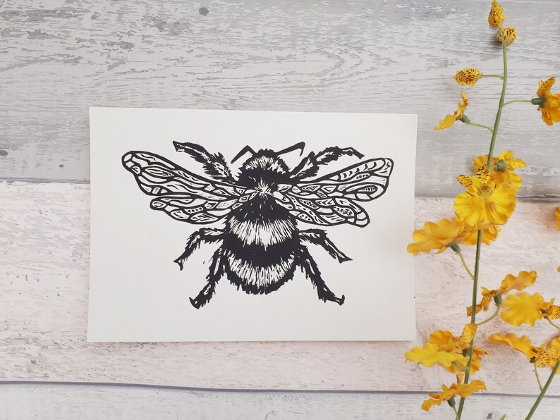Puede incluir: Una tarjeta rectangular blanca que presenta una impresi&oacute;n en bloque de tinta negra de un abejorro. La abeja est&aacute; representada con alas detalladas y estampadas, y un cuerpo difuso y texturizado. La ilustraci&oacute;n muestra l&iacute;neas negras de alto contraste.