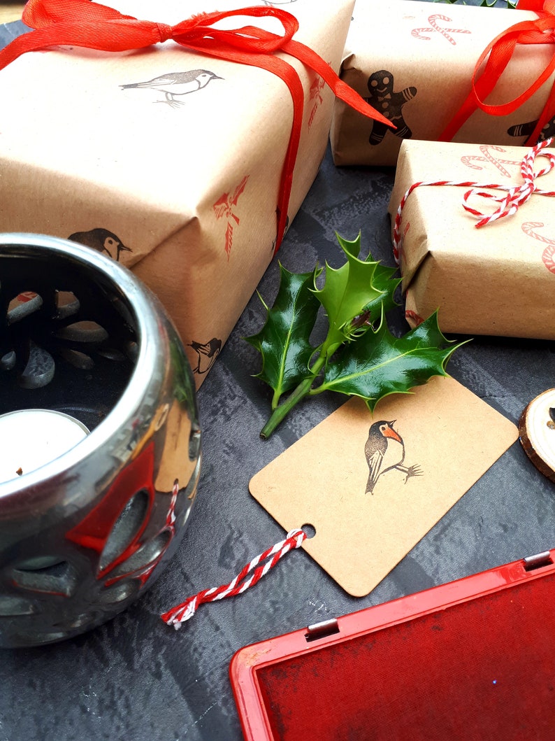 May include: Wrapped gifts with red ribbon and bird stamps. A silver candle holder, holly, and a red ink pad are also present. The gifts are wrapped in brown paper and tied with red ribbon.