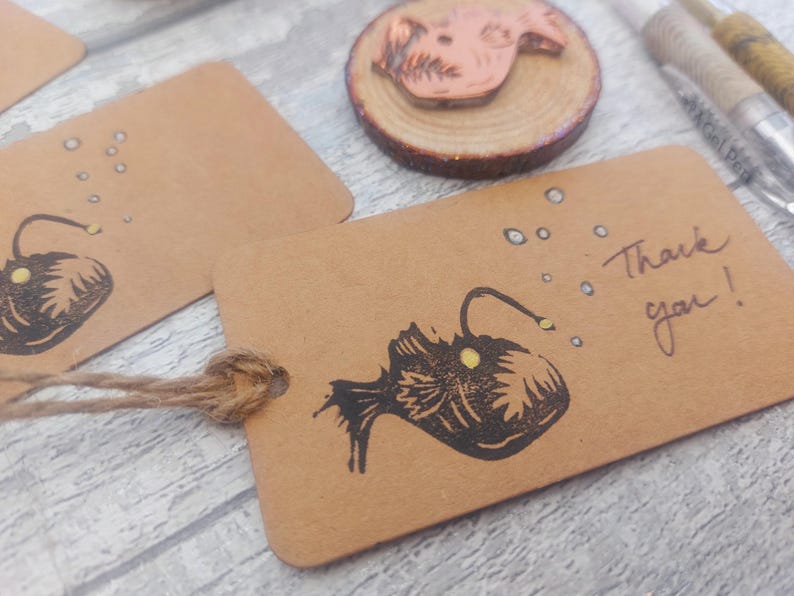 May include: A brown card with a black and white illustration of a anglerfish with a yellow lure. The card has the text "Thank you!" written on it.