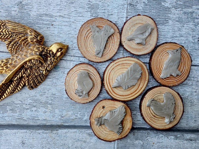 May include: A golden bird sculpture with outstretched wings is positioned near several small, round wooden slices. Each slice features a gray bird or feather design. The wood slices have visible growth rings, creating a rustic aesthetic.