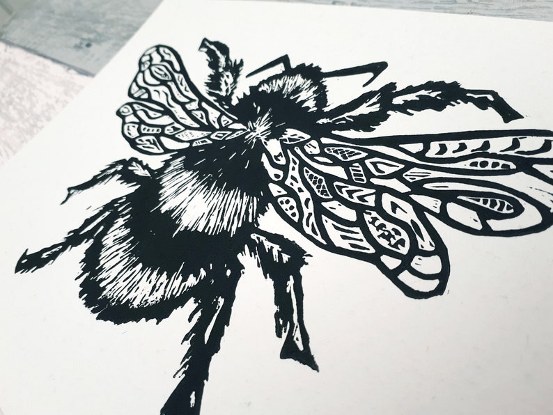 Puede incluir: Un grabado en lin&oacute;leo en blanco y negro sobre papel que muestra un abejorro detallado. El insecto est&aacute; representado con tinta negra de alto contraste, mostrando intrincados patrones geom&eacute;tricos en las alas y l&iacute;neas texturizadas y peludas.