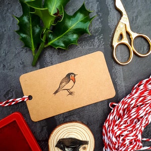 May include: A craft scene with a brown tag featuring a robin illustration, a wooden stamp, holly leaves, gold scissors, red ink pad, and red and white twine. The robin stamp is on a wood slice. The background is dark gray.