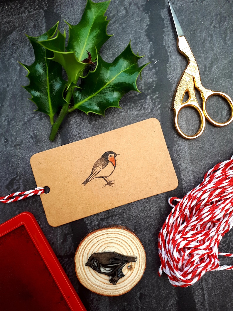 May include: A craft scene with a brown tag featuring a robin illustration, a wooden stamp, holly leaves, gold scissors, red ink pad, and red and white twine. The robin stamp is on a wood slice. The background is dark gray.