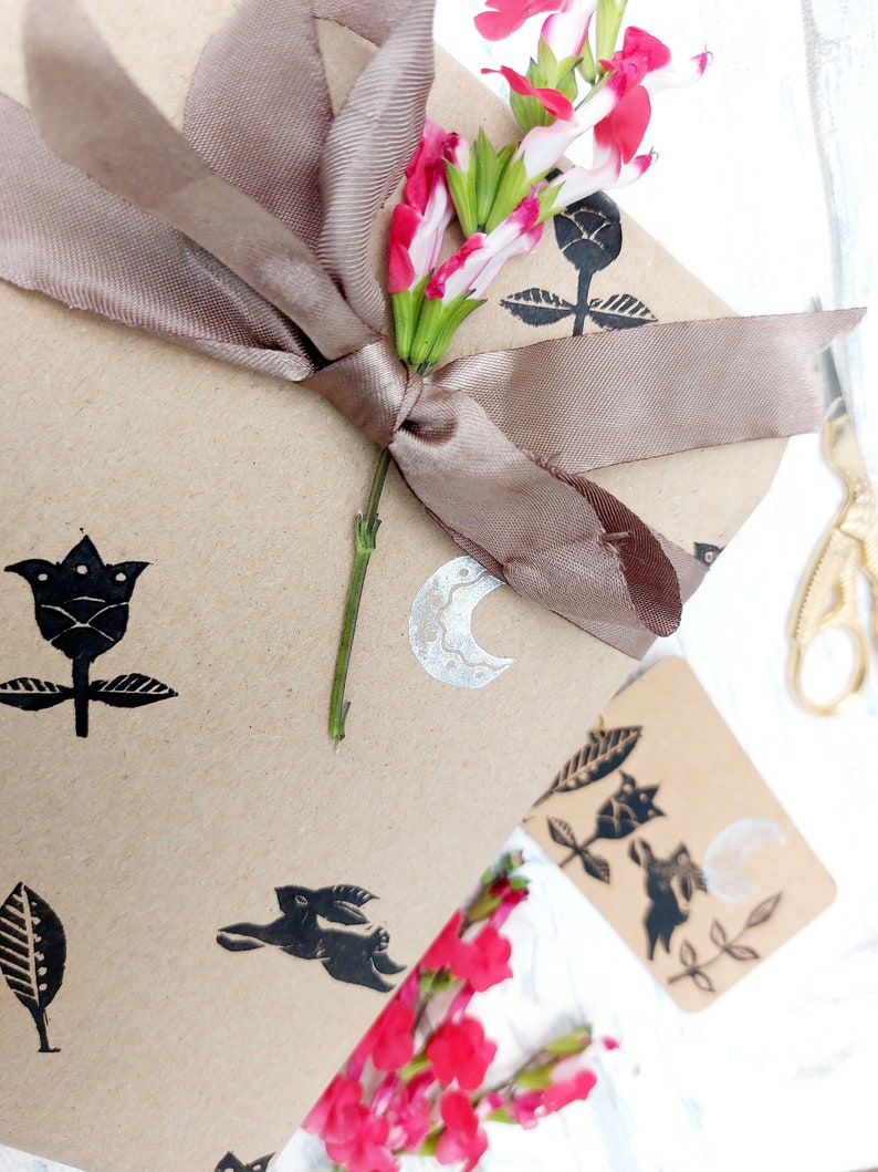May include: A brown paper gift bag with a brown satin ribbon tied around it. The bag is decorated with black and white stamps of flowers, leaves, a crescent moon, and a rabbit. A bouquet of pink and white flowers is on top of the bag.
