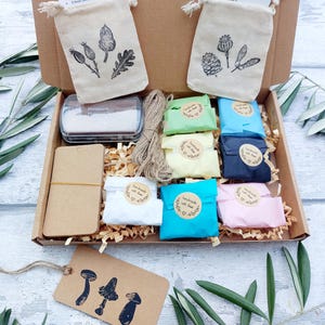 May include: A craft kit in a cardboard box, containing small fabric bags with botanical illustrations, colorful wrapped items, a stamp pad, twine, and tags with mushroom designs. The kit includes a variety of craft supplies.