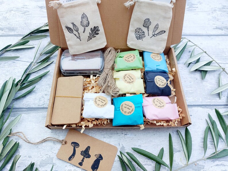 May include: A craft kit in a cardboard box, containing small fabric bags with botanical illustrations, colorful wrapped items, a stamp pad, twine, and tags with mushroom designs. The kit includes a variety of craft supplies.