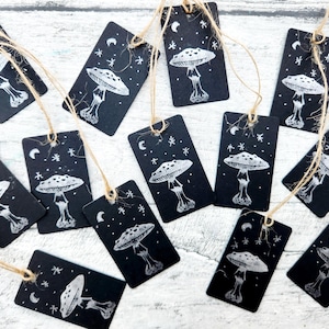 May include: A collection of black rectangular gift tags with a white illustration of a mushroom, moon, and stars. Each tag is strung with natural twine. The tags are arranged on a light-colored wooden surface.