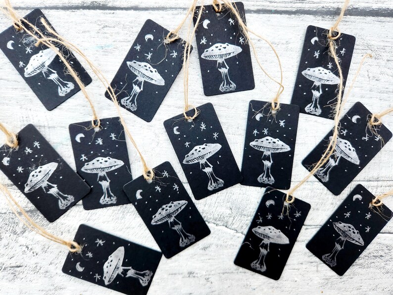 May include: A collection of black rectangular gift tags with a white illustration of a mushroom, moon, and stars. Each tag is strung with natural twine. The tags are arranged on a light-colored wooden surface.