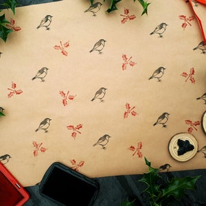 May include: A craft scene featuring brown wrapping paper printed with bird and holly designs. Gold scissors, a red ink pad, and stamps are visible. Green holly and a red ribbon add festive accents.