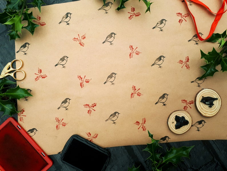May include: A craft scene featuring brown wrapping paper printed with bird and holly designs. Gold scissors, a red ink pad, and stamps are visible. Green holly and a red ribbon add festive accents.