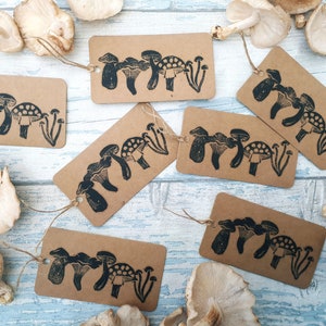 May include: Kraft paper gift tags with a black mushroom design and twine. The tags are rectangular and feature various mushroom illustrations. Several fresh mushrooms are scattered around the tags on a light blue wooden surface.